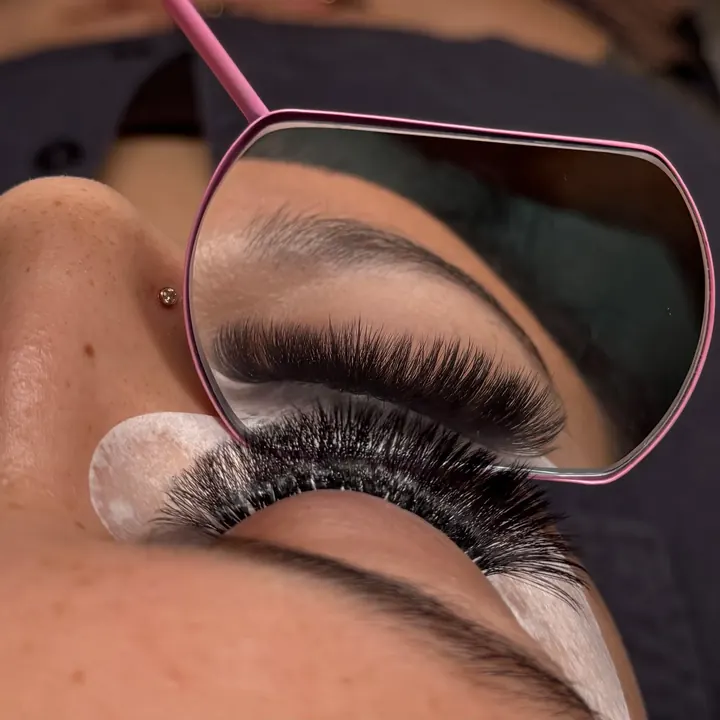 A client reclining with eyes closed while a pink hand mirror shows her new full, fluffy lash set
